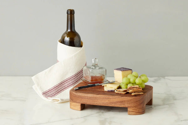Wooden cheese board with wine, cheese, square jam jar, and grapes on a marble surface.