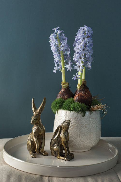 White ceramic pot with purple flowers and greenery, accompanied by two small bronze rabbit sculptures on a dark background.