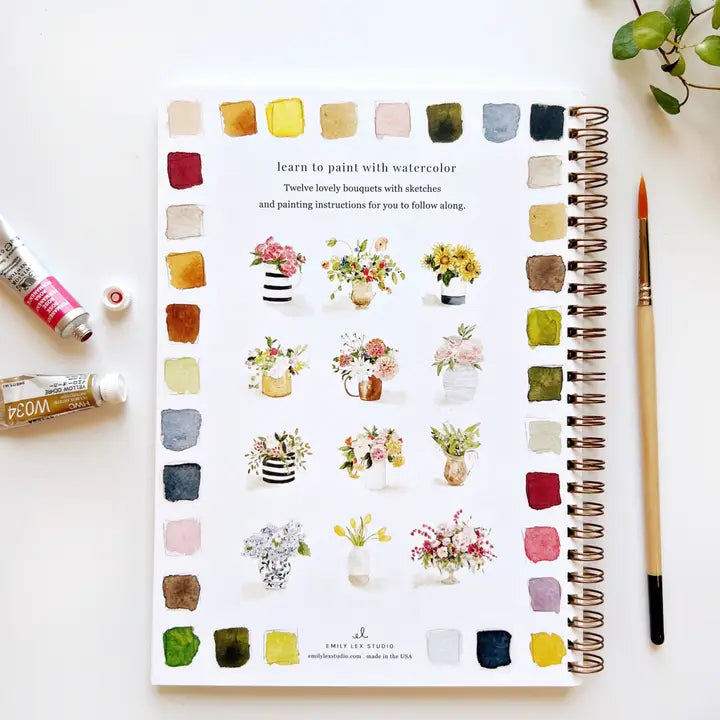 Book about watercolor painting with color swatches and flower illustrations, surrounded by art supplies.