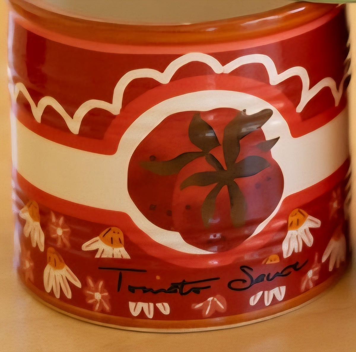A close-up of a red ceramic vase painted with 'Tomato Sauce' with a tomato illustration and small floral motifs on a beige surface.