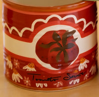A close-up of a red ceramic vase painted with 'Tomato Sauce' with a tomato illustration and small floral motifs on a beige surface.