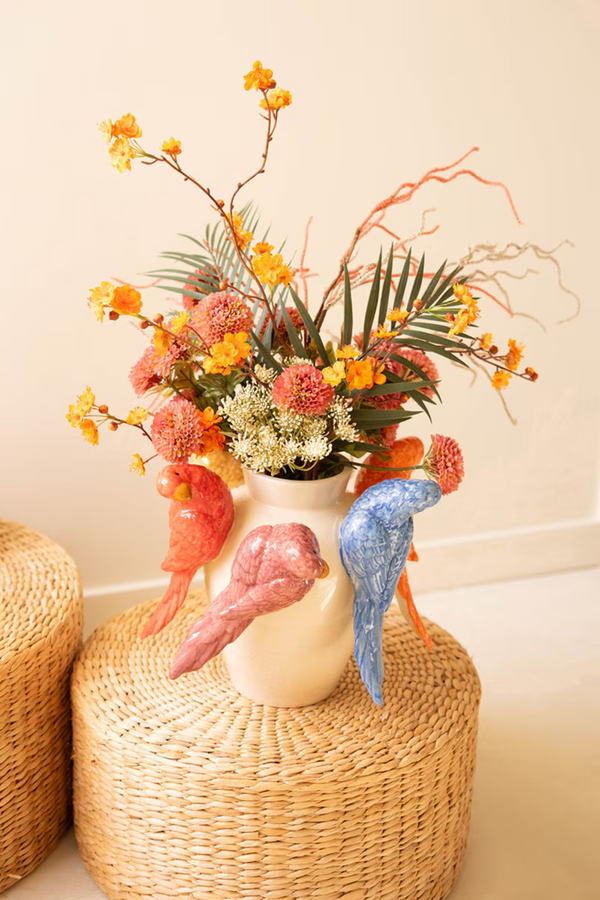 Decorative vase featuring parrots on the sides and filled with flowers on a woven stool against a plain wall