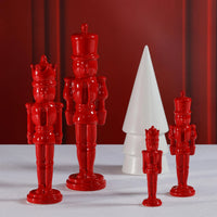 Four glossy red nutcracker figurines in different sizes arranged on a white surface with a red draped backdrop, alongside a white cone-shaped decorative object.