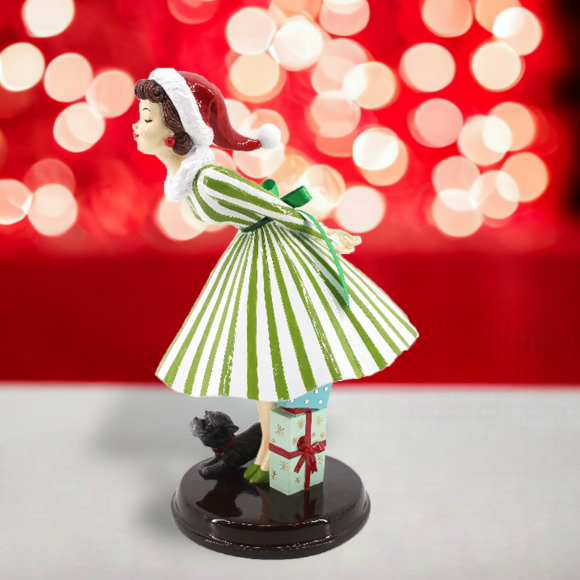 Figurine of a woman in a green and white striped dress and a Santa hat leaning over for a kiss with a dog, against a blurred red and white light background.