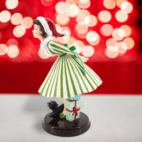 Figurine of a woman in a green and white striped dress and a Santa hat leaning over for a kiss with a dog, against a blurred red and white light background.