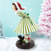 Christmas-themed figurine of a woman in a green dress with a Santa hat, standing next to a pink Christmas tree.