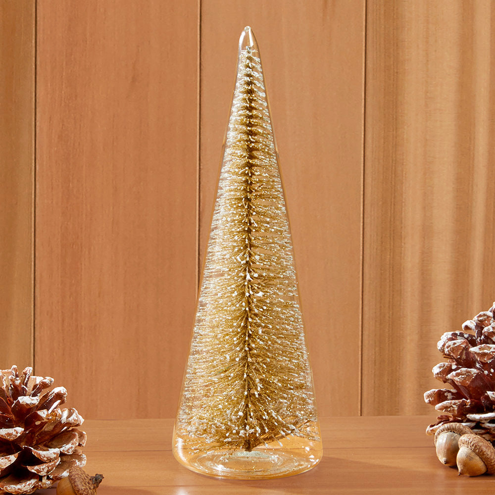 A tall, slender gold glitter Christmas tree decoration inside of glass, standing on a wooden surface, with pinecones on both sides against a wood-paneled background.