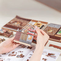 Hand holding a miniature model of a building with a detailed interior, against a background of similar models and a manual.