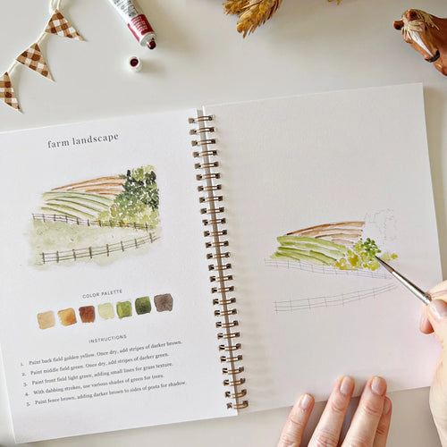 Artistic watercolor workbook with a person painting a farm landscape and color palette on a light surface.