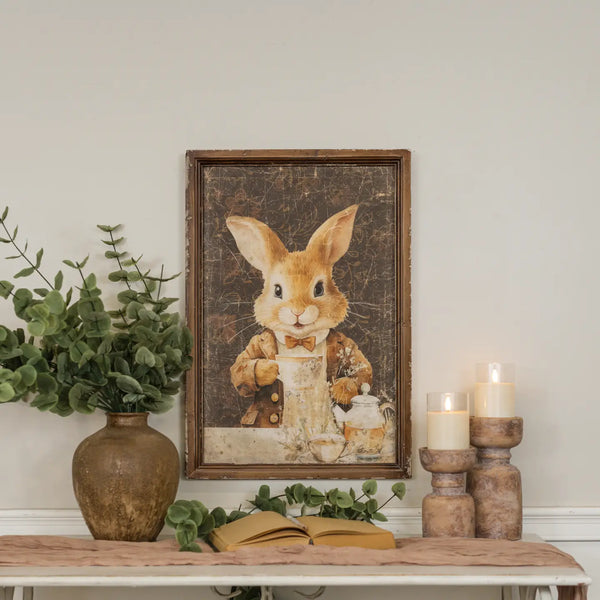 Decorative setup with a framed picture of a rabbit, candles, and plants on a table.