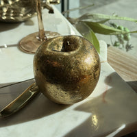 A decorative metallic gold apple sculpture with a distressed, textured finish, styled on a white marble tray with a gold knife and glass base in the background.