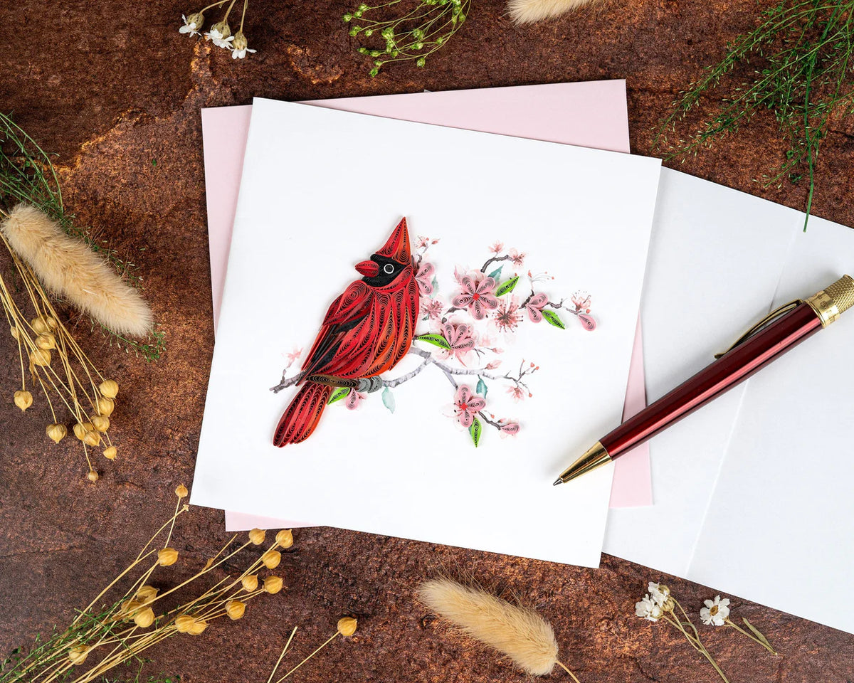 Card with a quilled red cardinal illustration on a rustic surface with a pen and flowers.