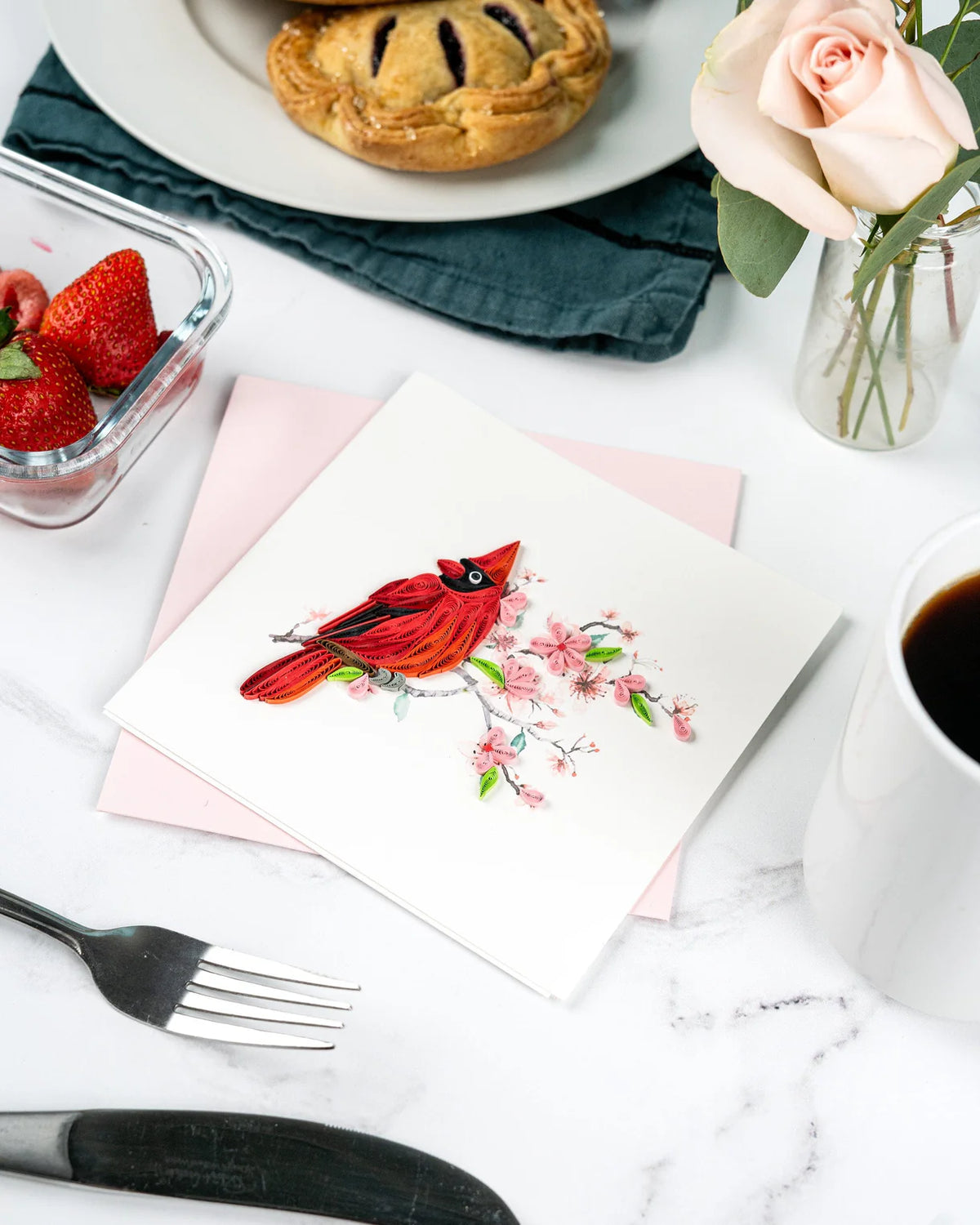 Card with quilled red cardinal and cherry blossoms on a table with food and coffee.