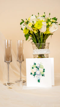 Two champagne flutes, a vase of flowers, and a wedding card on a beige surface.