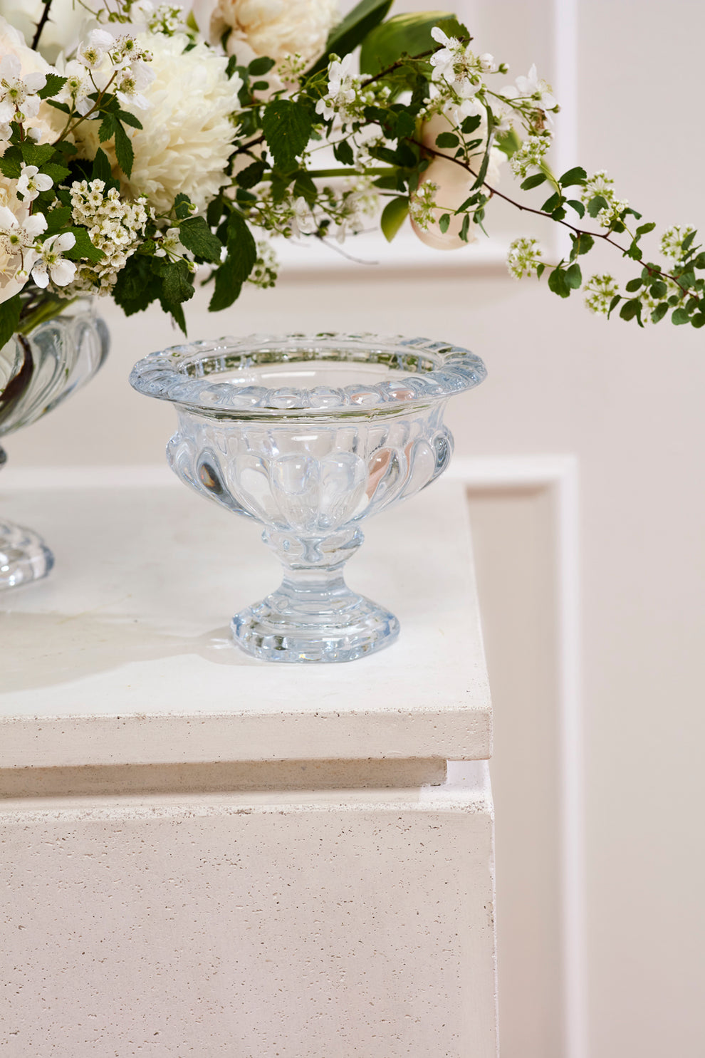 A clear glass pedestal bowl with a scalloped rim and footed base, styled on a white textured pedestal with white flowers in the background.