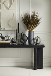 Decorative setup with a vase of dried grasses and raven figurines on a black console table against a white paneled wall.