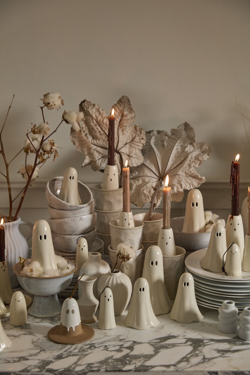 A Halloween decor display featuring ghost figurines and ghost candleholders with lit candles, in gray-white bowls and plates, with faux leaves and cotton, all on a marble surface.