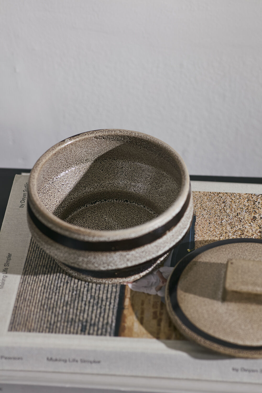 A speckled ceramic container with a matching lid, styled on top of a newspaper against a neutral wall.