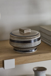A speckled ceramic lidded box with dark brown stripes sits on a light wood shelf in a minimalist interior setting.
