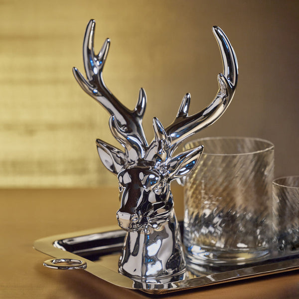 A polished silver stag head decorative object with antlers, shown on a metal tray beside two clear drinking glasses against a warm beige background.