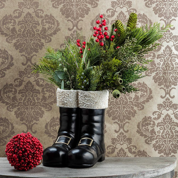 A vase made to look like Santa's boots with evergreens and berry sprays inside on a gray wooden table next to a berry bunch and a wallpapered wall in the background.