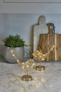 Decorative gold bird sculptures with LED lights on a marble surface, with a plant and cutting board in the background.