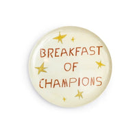 A round, glossy cream-colored melamine plate featuring the text 'BREAKFAST OF CHAMPIONS' in a rustic brown font, decorated with small gold stars, shown on a plain white background.