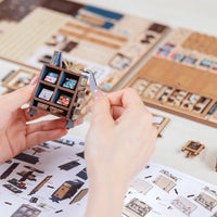 Person holding a small model of a room with a detailed layout, surrounded by similar models and design sheets.